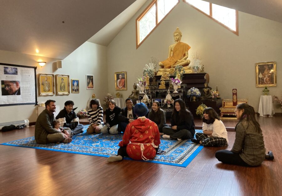 a group of students sits on the floor of a Buddhist temple with a "nunk" wearing a red cloak