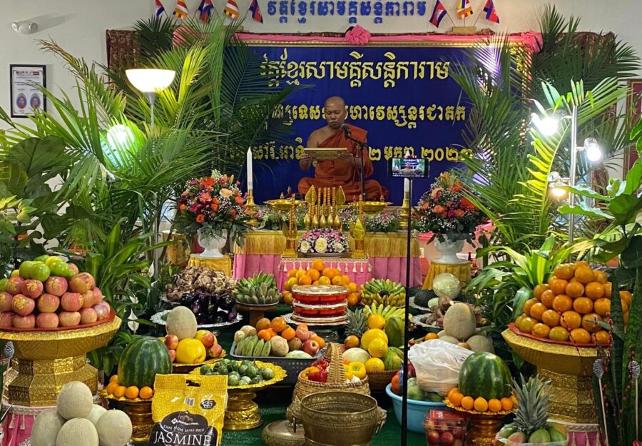 monk in saffron robe sits on altar surrounded by bowls of fruit and other offerings