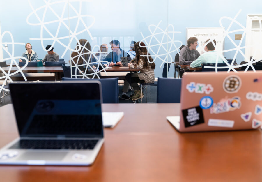 two laptops are open on a desk; a class of high school students on the other side of the glass