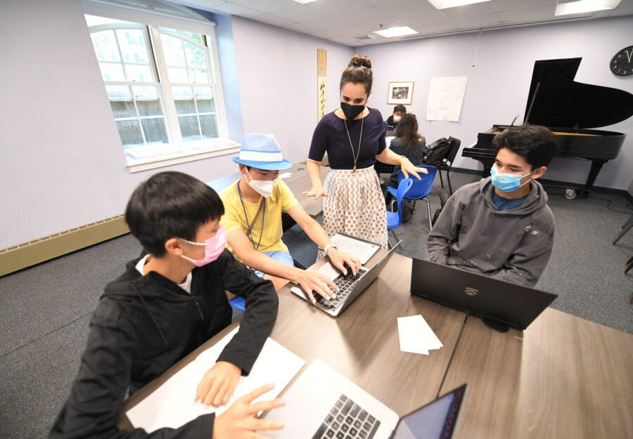 female instructor stands in front of three male students who are seated at a table; they are engaged in conversation