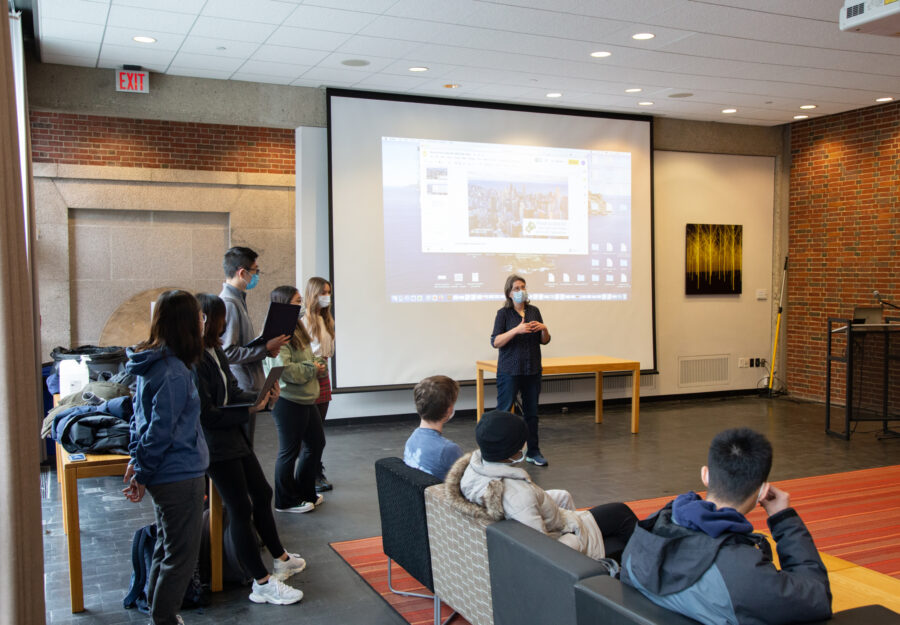 students gathered in large room with an orange rug and a white board; teacher talking in the front of the room
