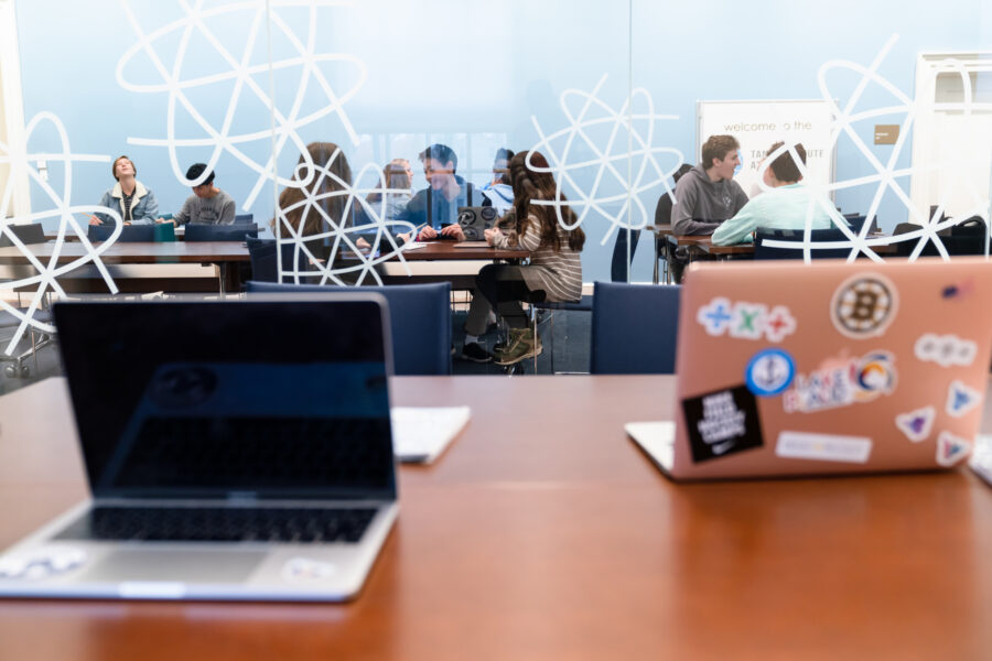 two laptops are open on a desk; a class of high school students on the other side of the glass