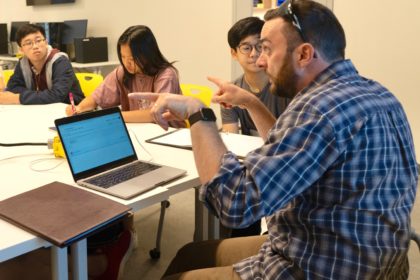 male teacher sitting at a table discussing ethics in a CS classroom with students