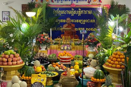monk in saffron robe sits on altar surrounded by bowls of fruit and other offerings