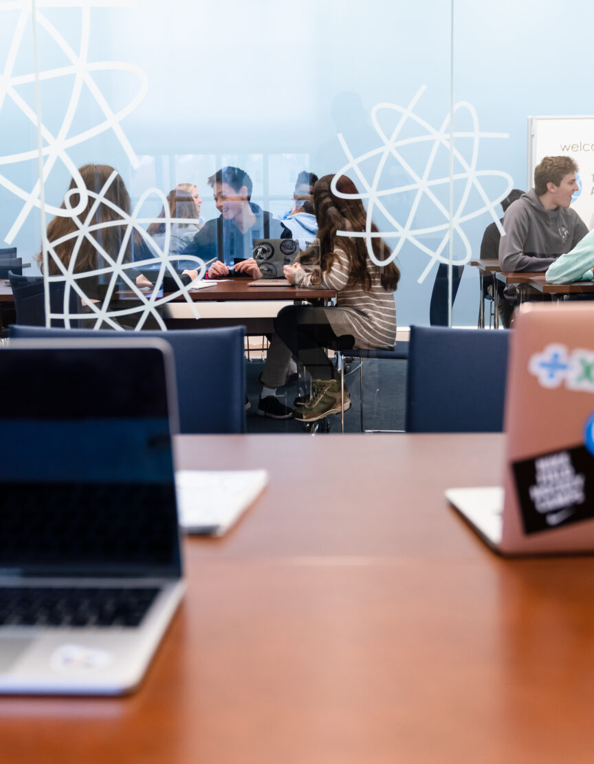 two laptops are open on a desk; a class of high school students on the other side of the glass