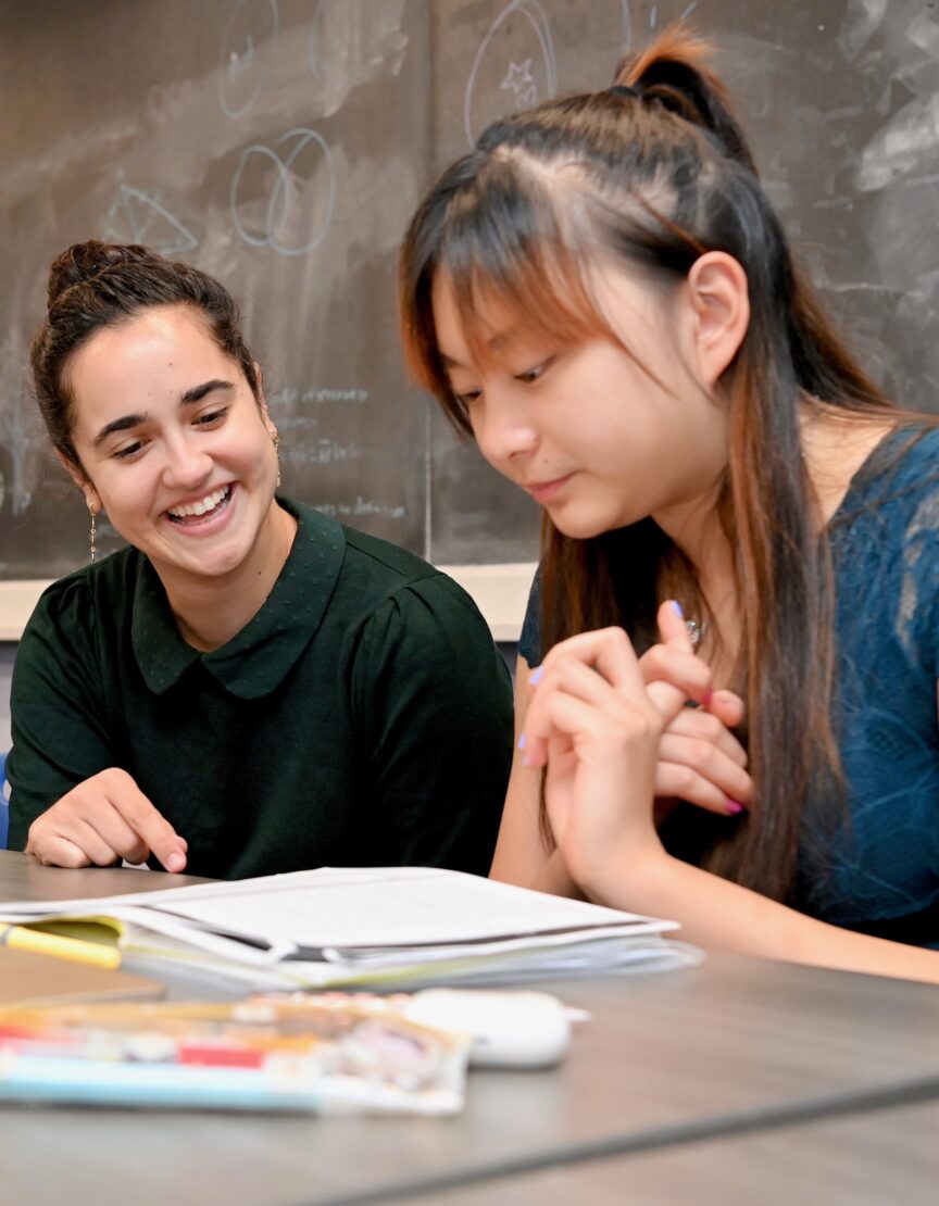 female teacher sitting at a table with a student; she is smiling; there is a notebook open on the table