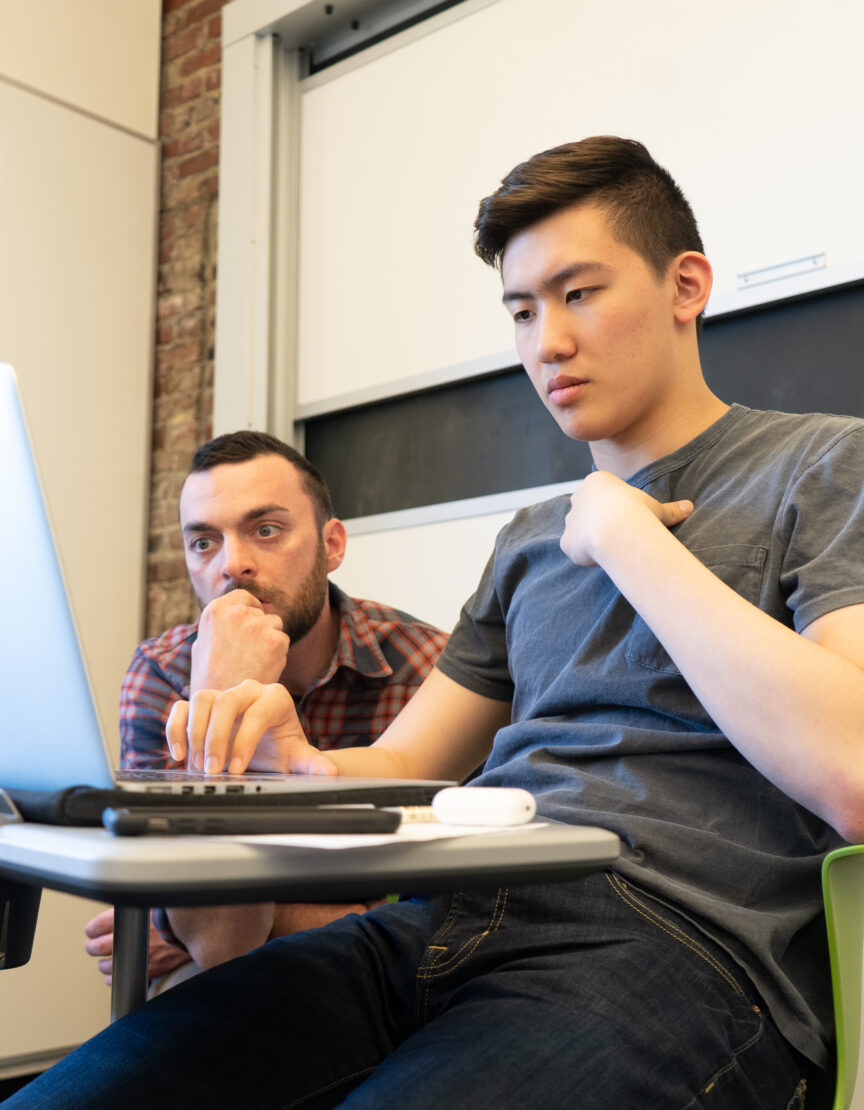 male student sitting at a desk typing on a laptop with male instructor looking over his shoulder