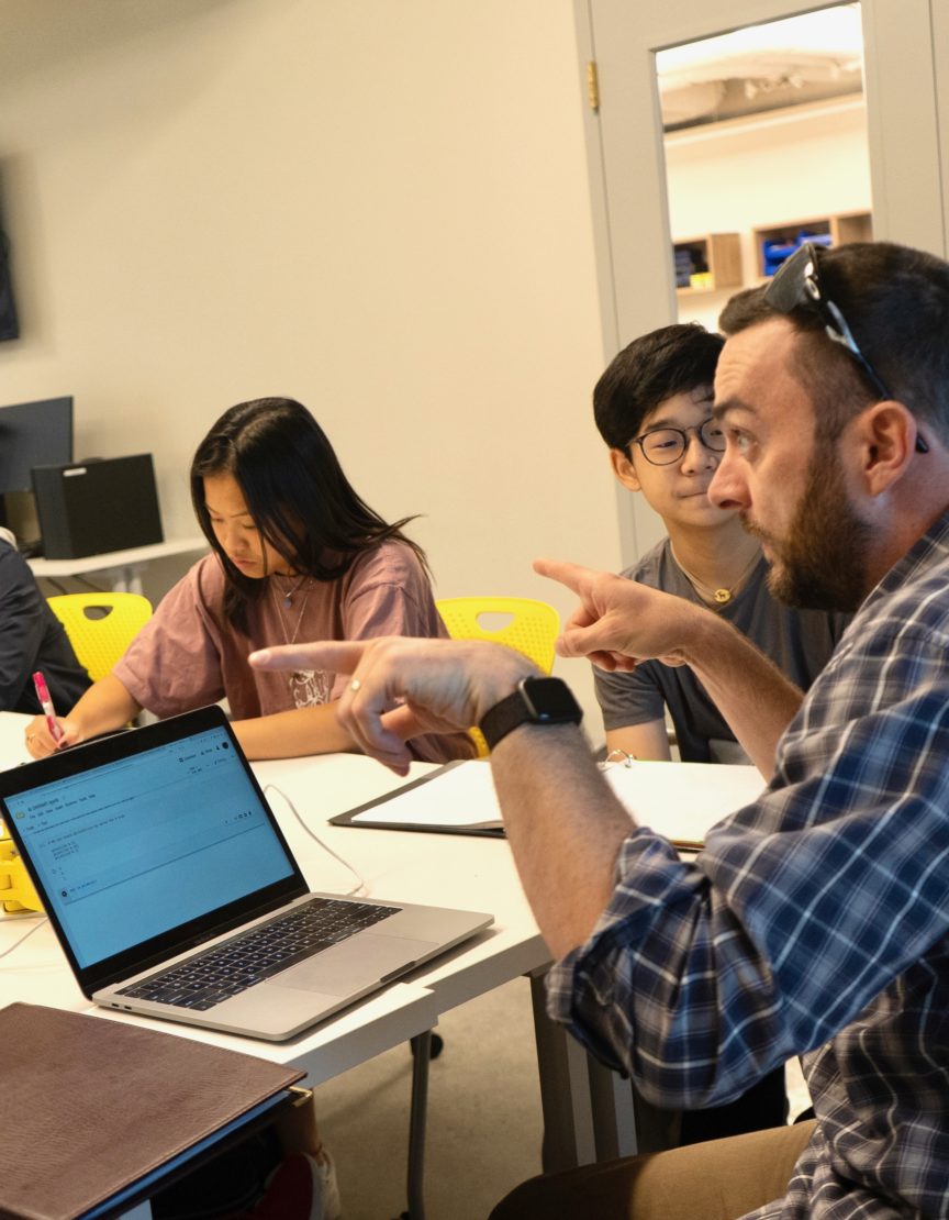 male teacher sitting at a table discussing ethics in a CS classroom with students