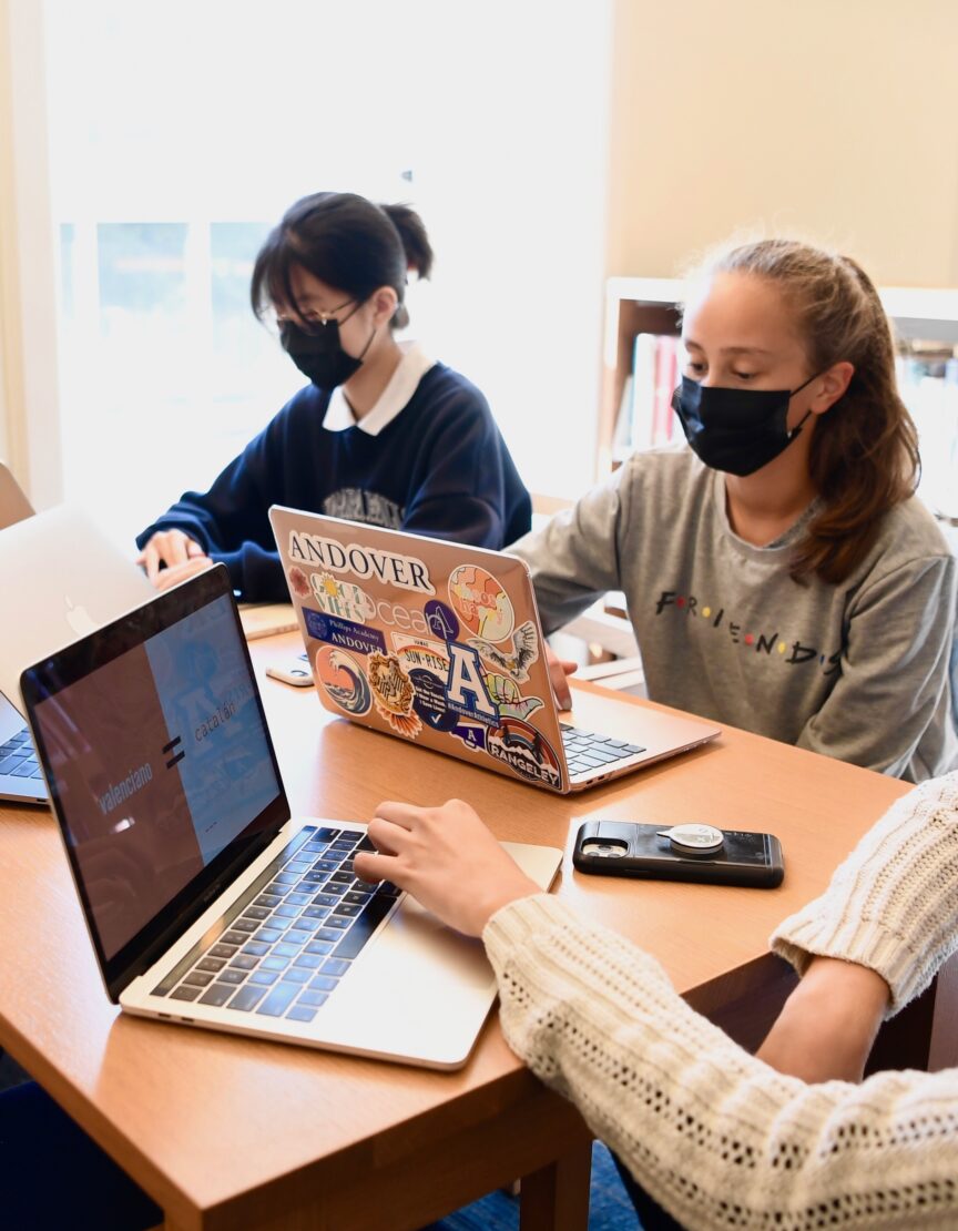 two girls sit at a table with laptops