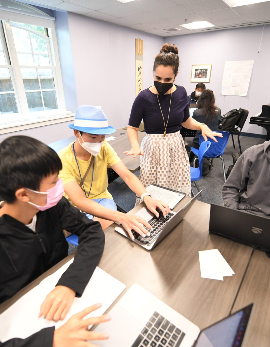 female instructor stands in front of three male students who are seated at a table; they are engaged in conversation