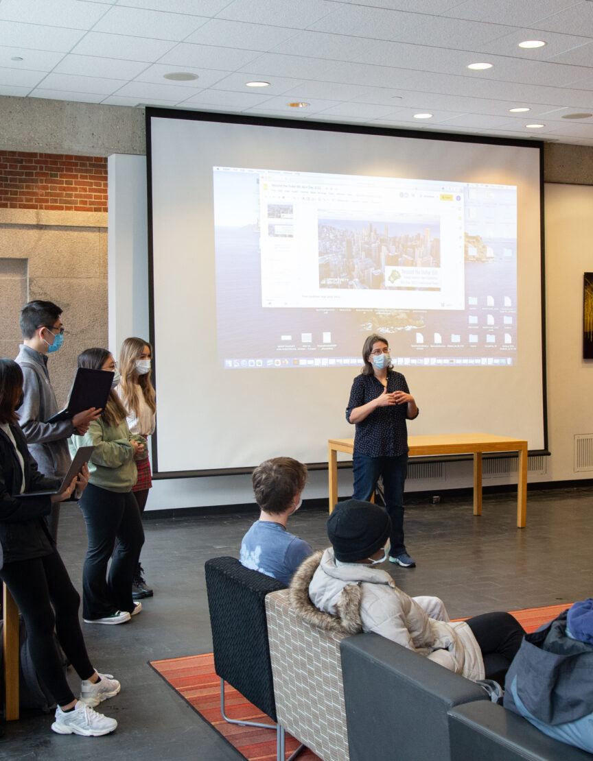 students gathered in large room with an orange rug and a white board; teacher talking in the front of the room